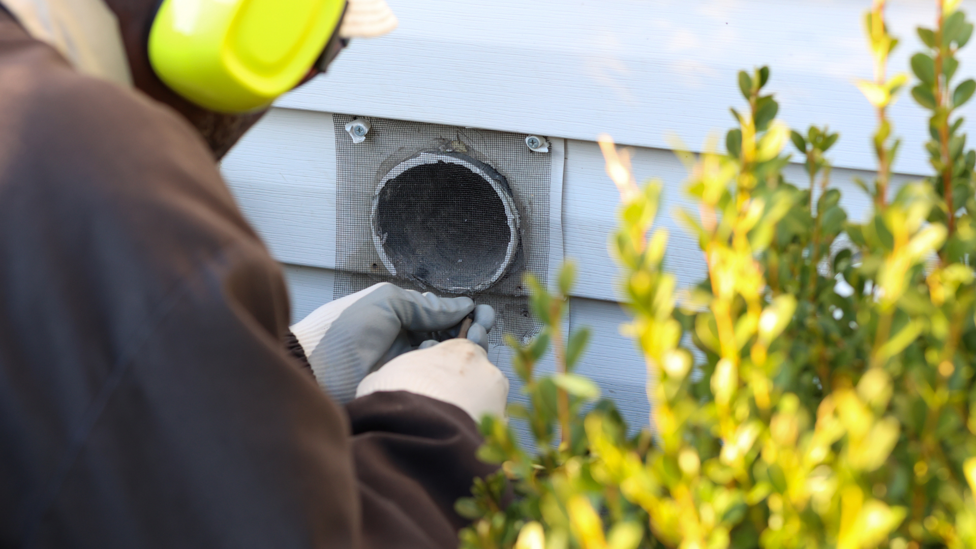 Person with gloves working on a circular vent on a building, wearing earmuffs.