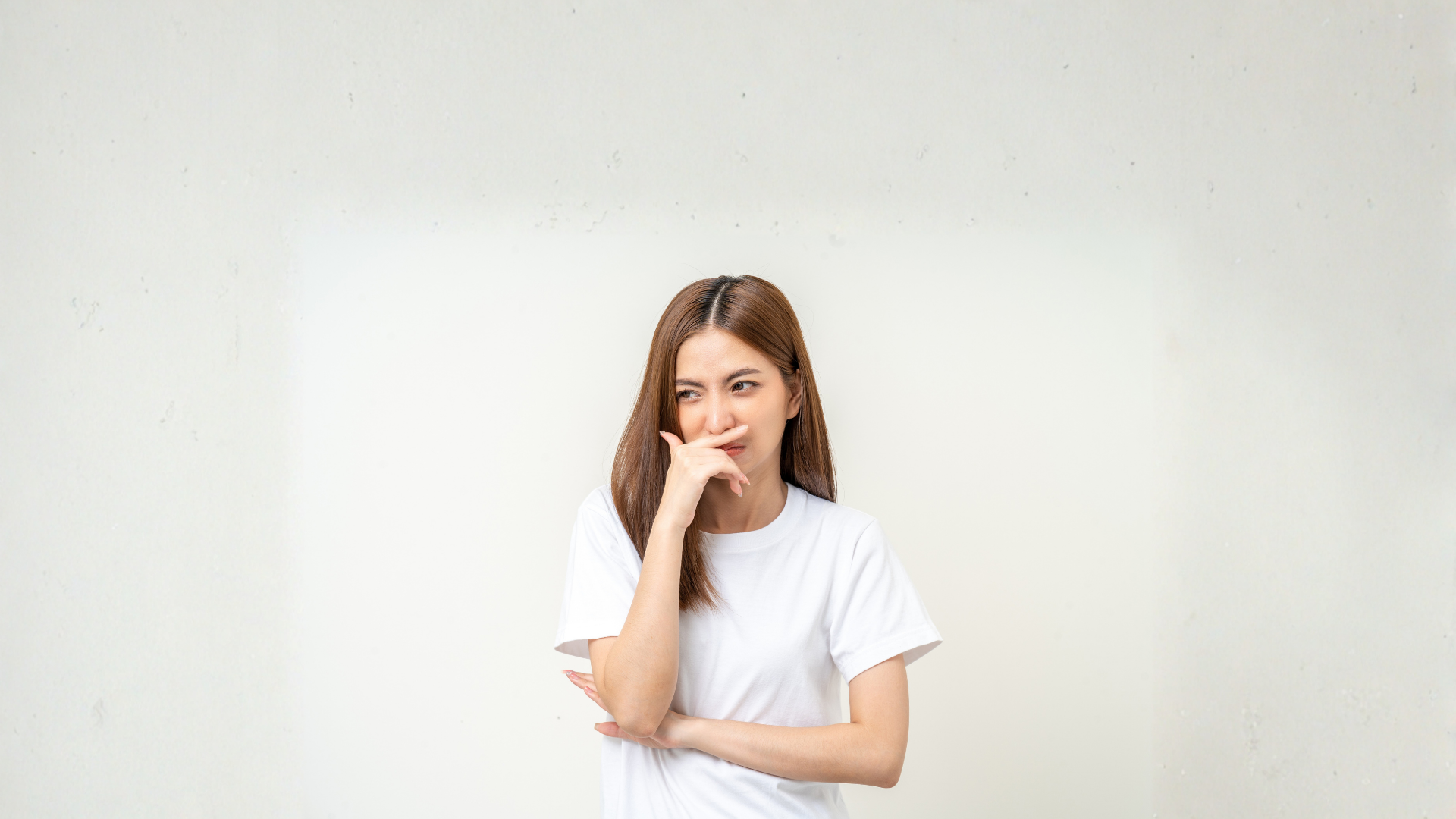 Woman in white t-shirt holding her nose, looking displeased. White wall background.