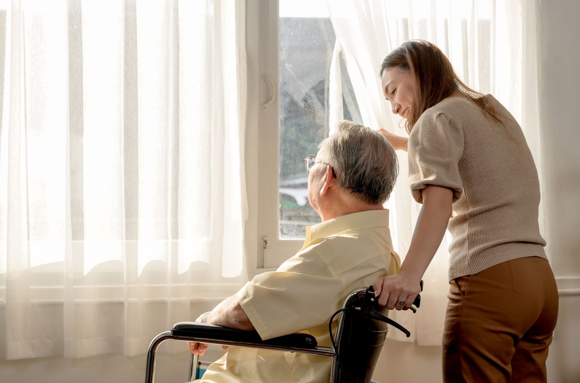 A woman is helping an elderly man in a wheelchair open a window.