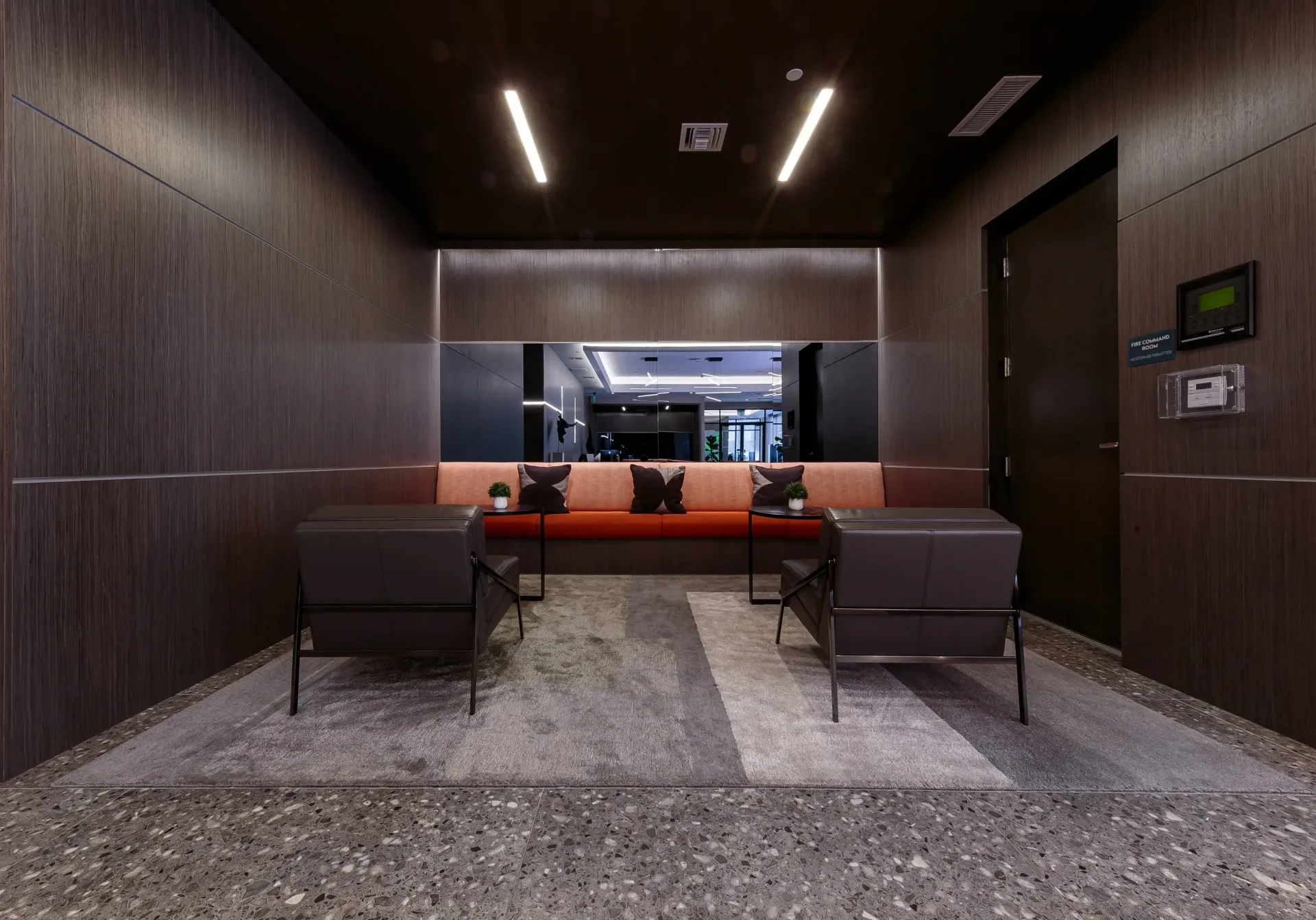 Modern apartment lobby with dark wood walls, a long orange sofa, two chairs, and small plants.