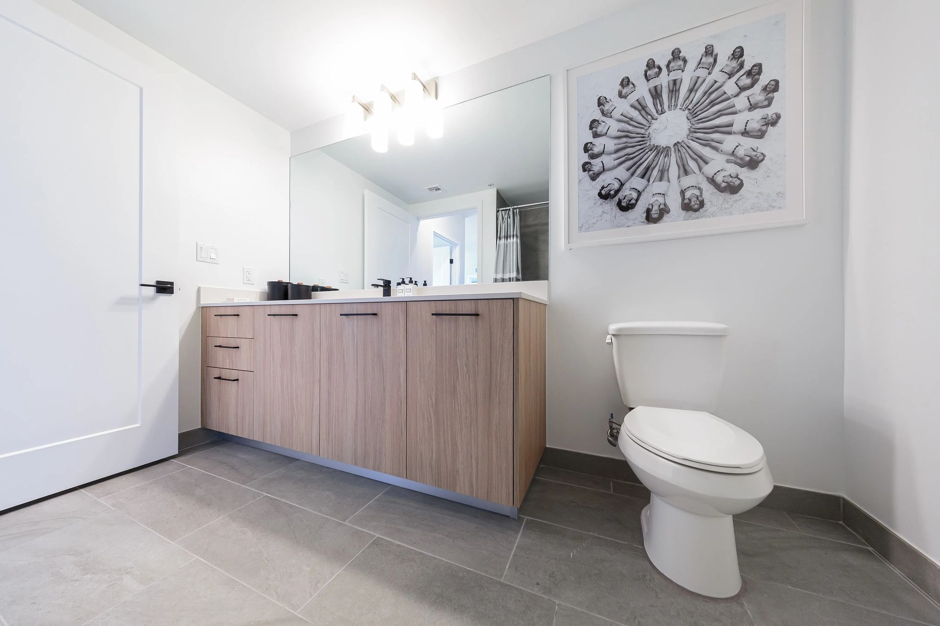 Bathroom with light wood vanity, white toilet, framed art, and gray tile flooring.