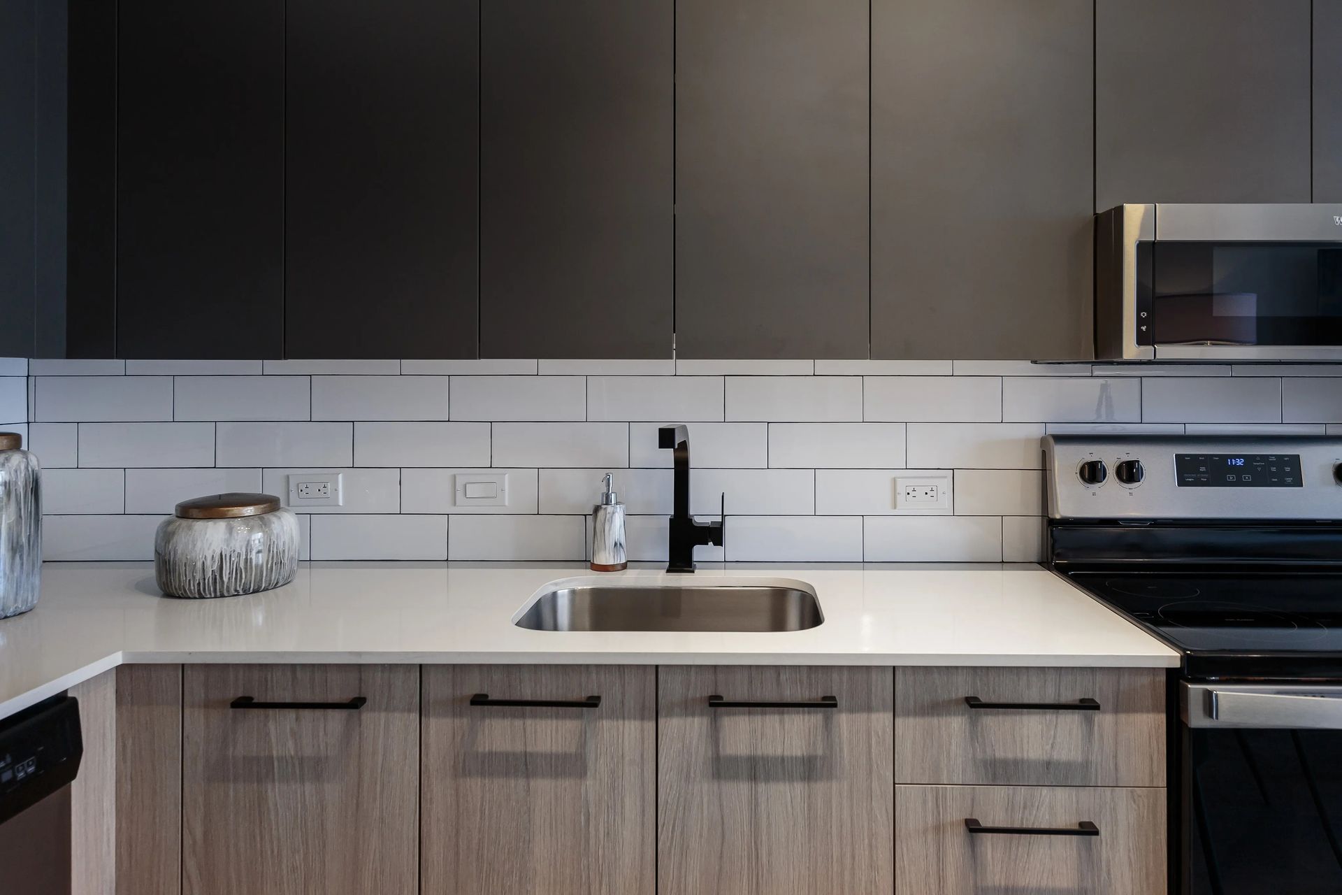 Kitchen with gray and light wood cabinets, white countertop, stainless steel sink, and black faucet.