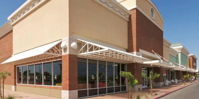 Strip mall storefront with tan and brick facade, glass windows, and white awnings on a sunny day