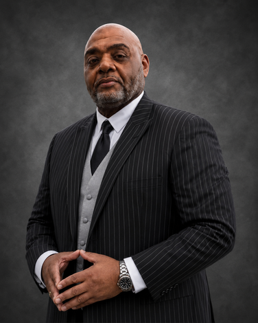 Man in a dark suit and bow tie, hands clasped, looking directly at the viewer.