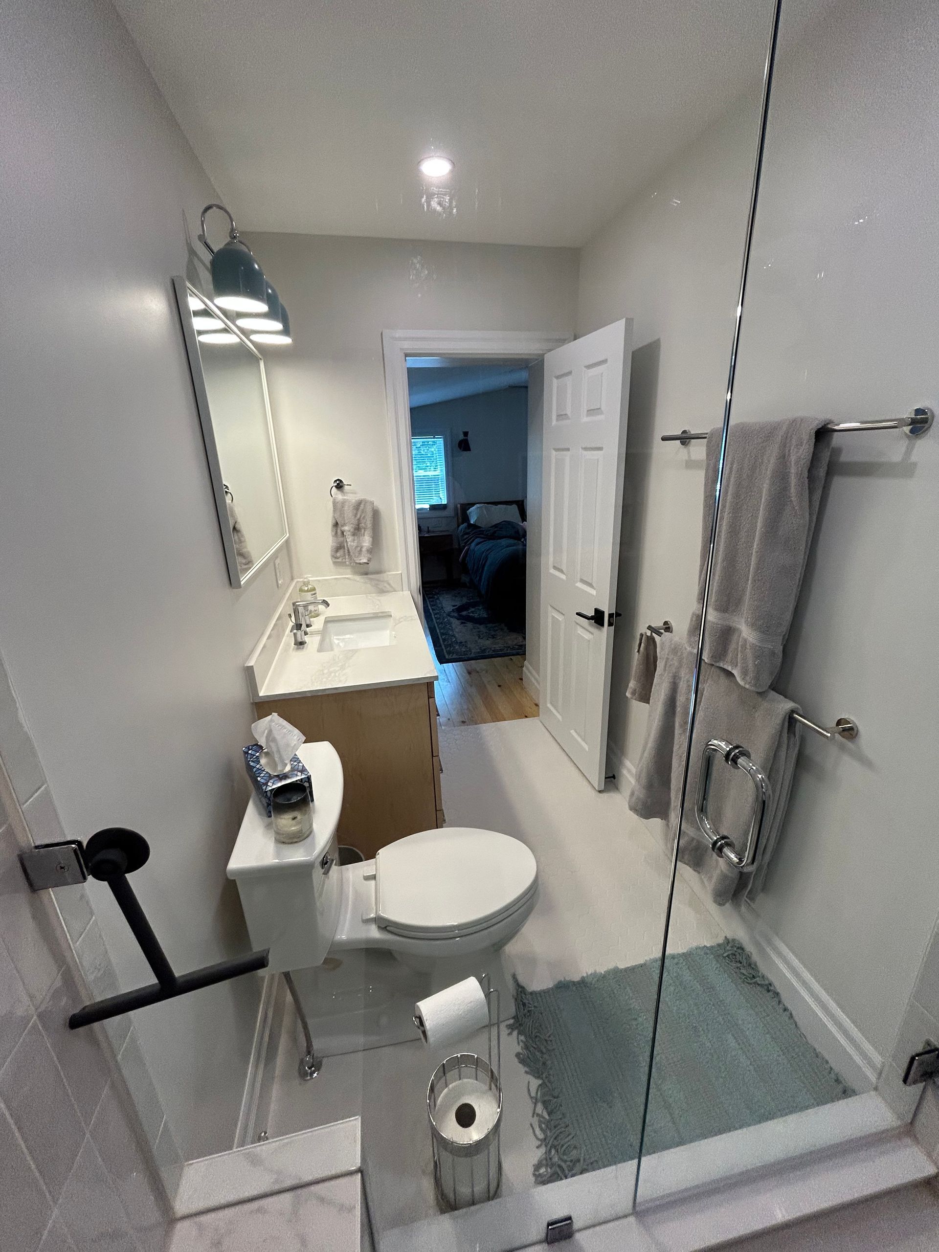 A bathroom with a toilet, a vanity sink, a glass shower door with towels on a rack, and a door leading to a bedroom.