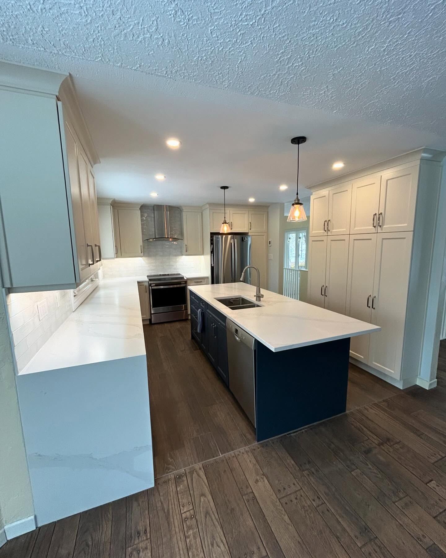 Modern kitchen with blue island, white countertops, stainless steel appliances, and wood flooring.