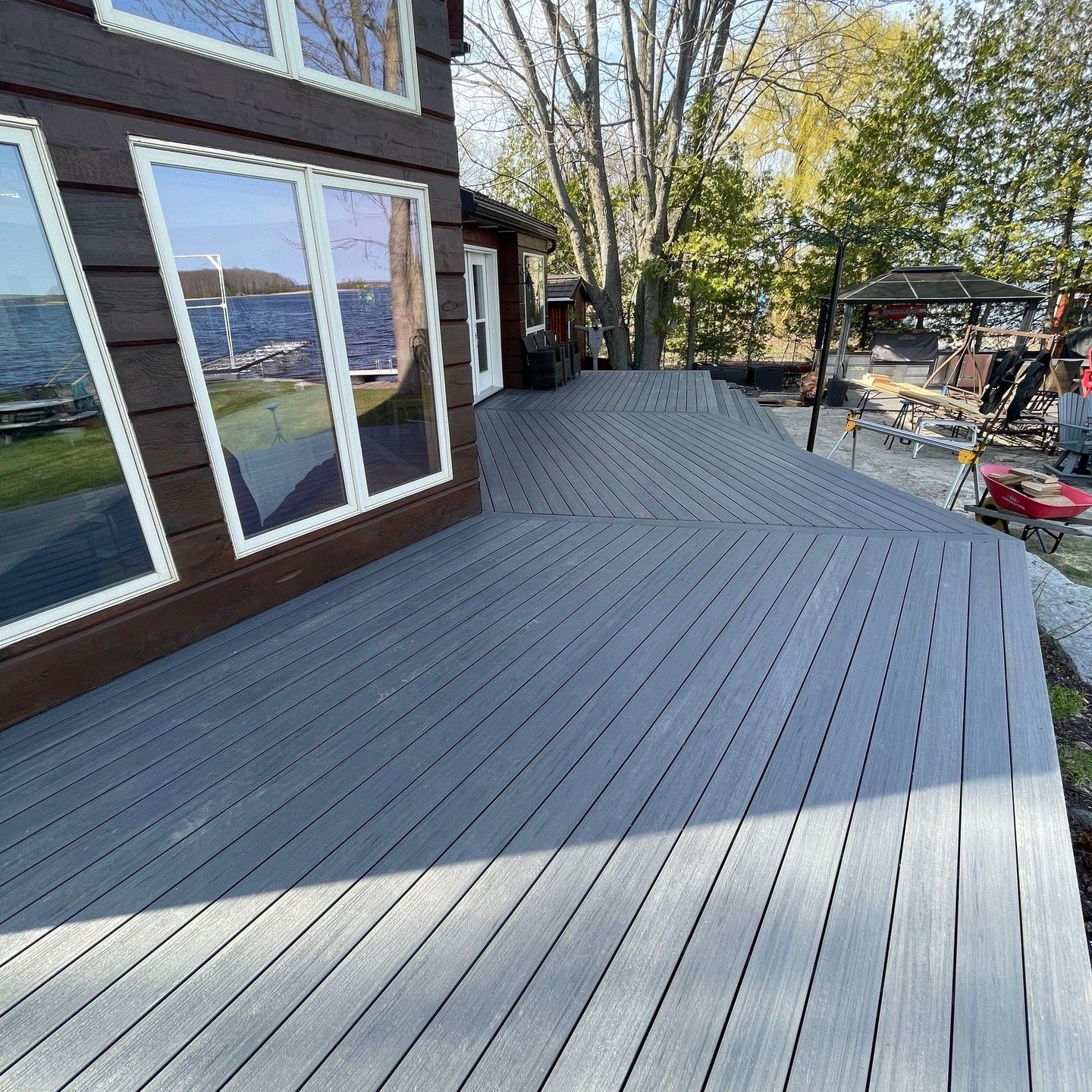 A gray composite deck extends from a brown house with large windows, overlooking a lake.