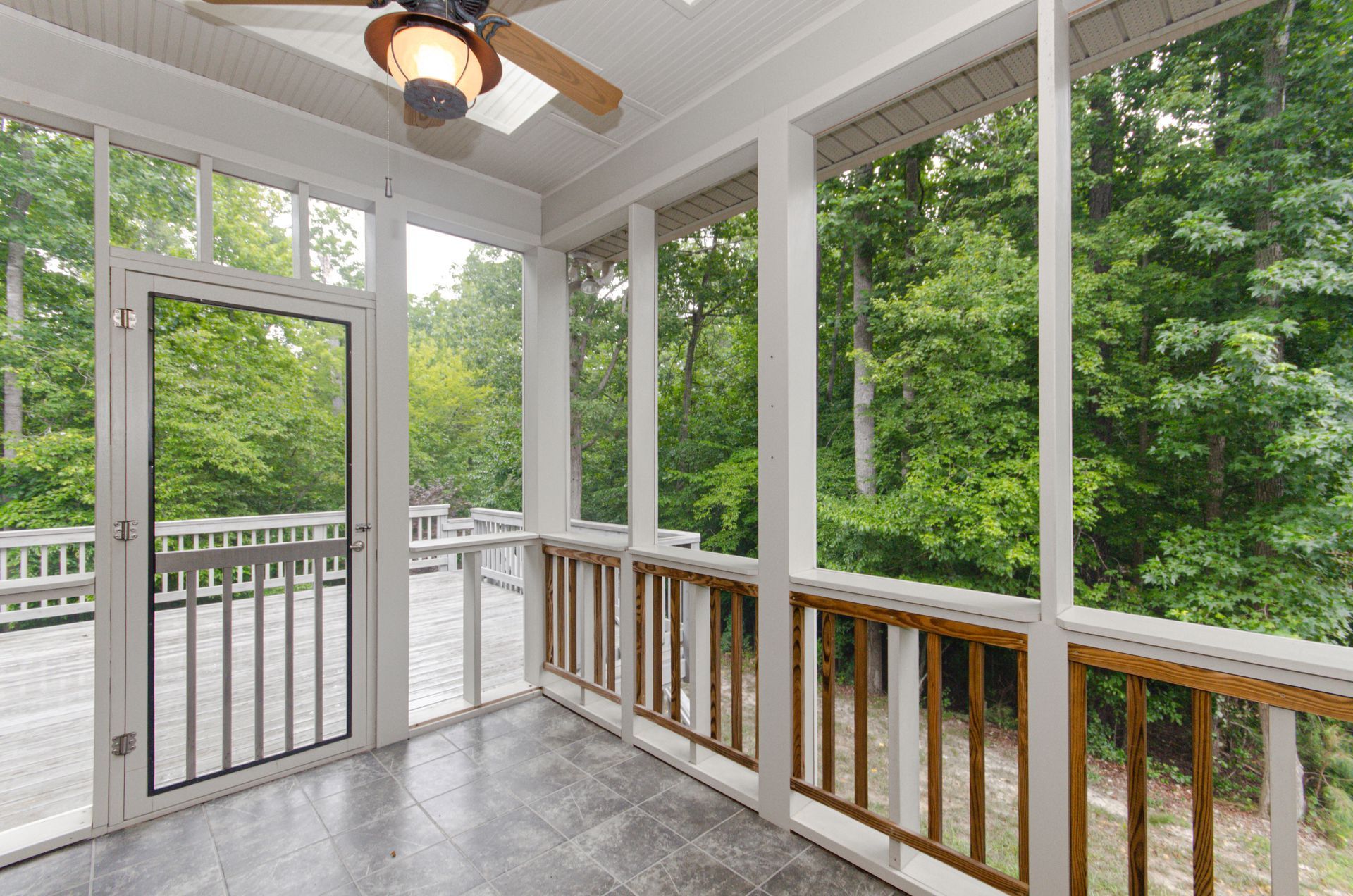 A screened-in porch with grey tile flooring, white framing, a ceiling fan, and a view of trees through the mesh walls.