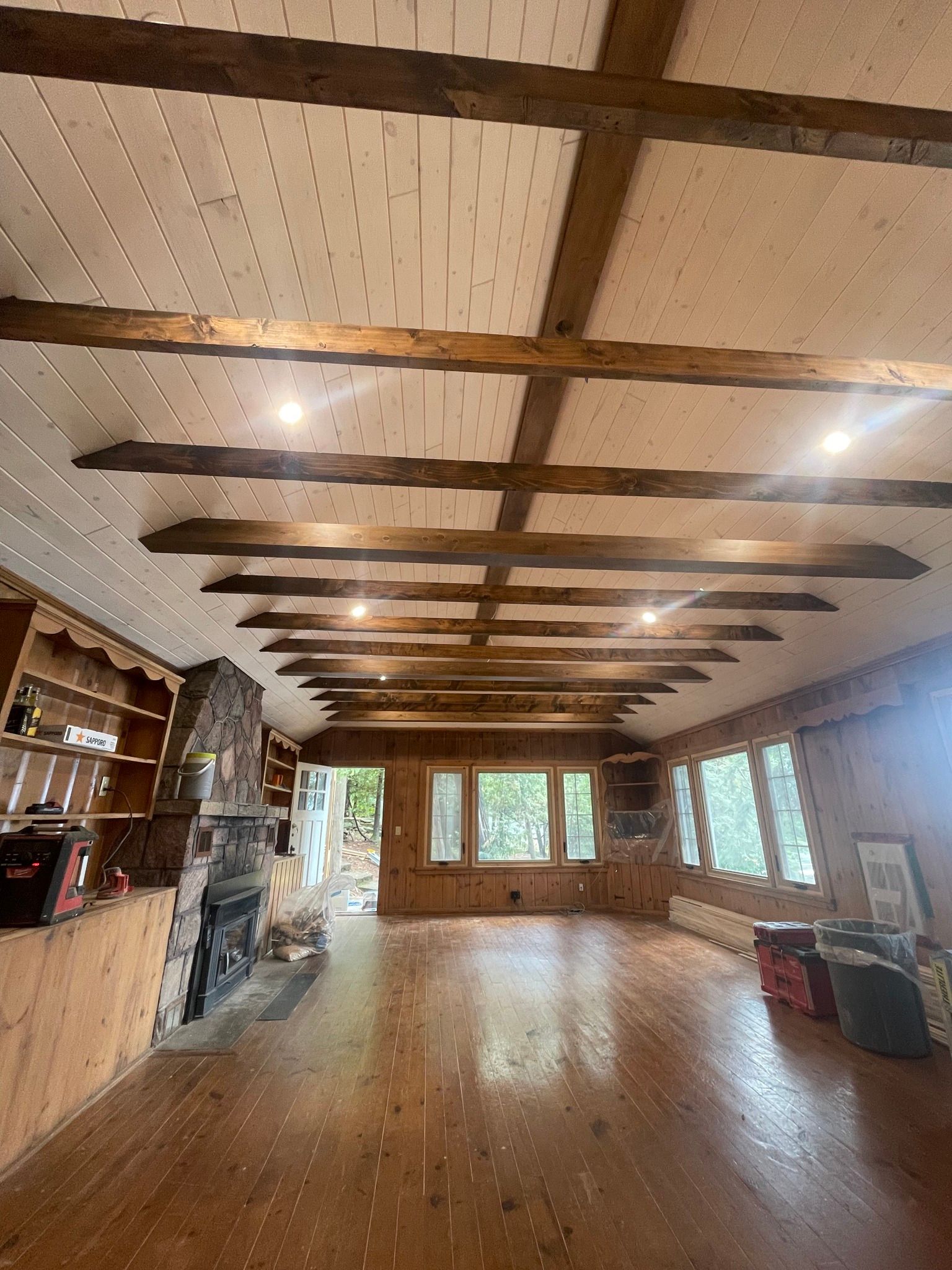 Spacious interior with exposed wooden beams, fireplace, and hardwood floors. Windows offer a view of the outdoors.