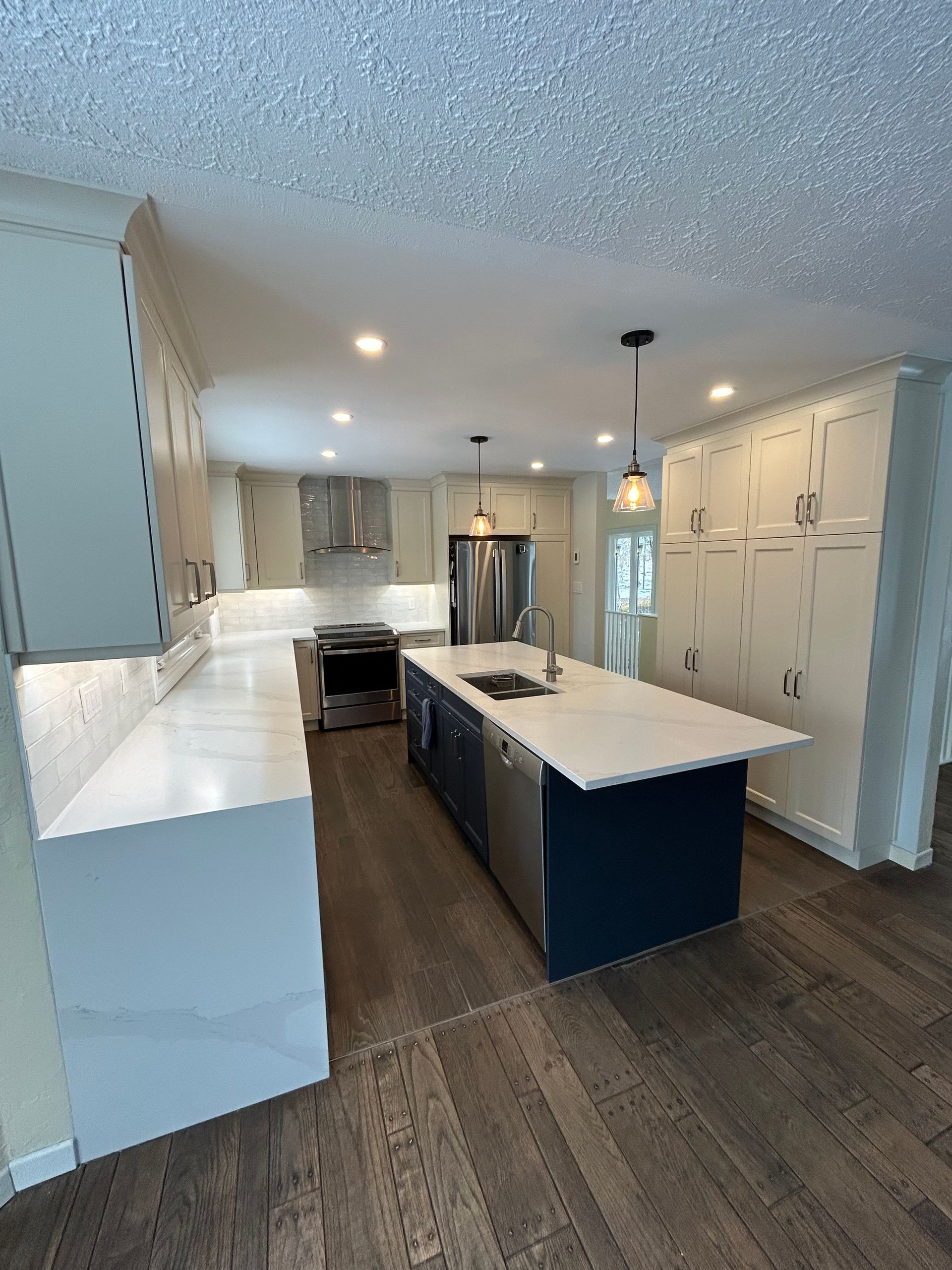Modern kitchen with white countertops, blue island cabinetry, light-colored tall cabinets, and wood-look flooring.