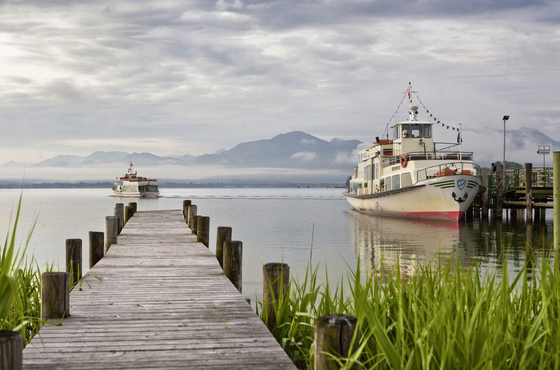 Wooden pier extends to a lake with boats, mountains in the background, overcast sky.
