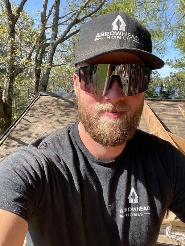 Man wearing sunglasses and black cap outdoors in sunlight, with trees and rooftops behind him