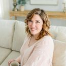 Woman with curly hair in pink sweater smiles while sitting on a couch.
