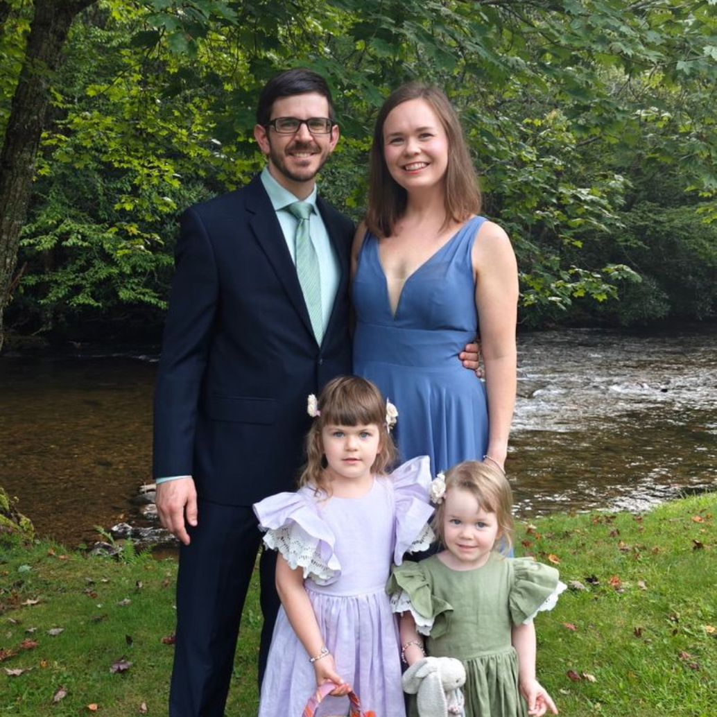 Family of four smiles for the camera by a river: man in a suit, woman in a blue dress, and two young girls in dresses.