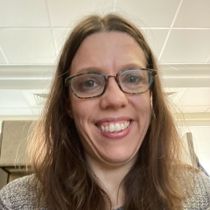 Woman with glasses smiles; long brown hair, grey sweater, indoors.