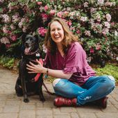 Woman smiling with black dog, sitting cross-legged on brick path, in front of pink flowers.