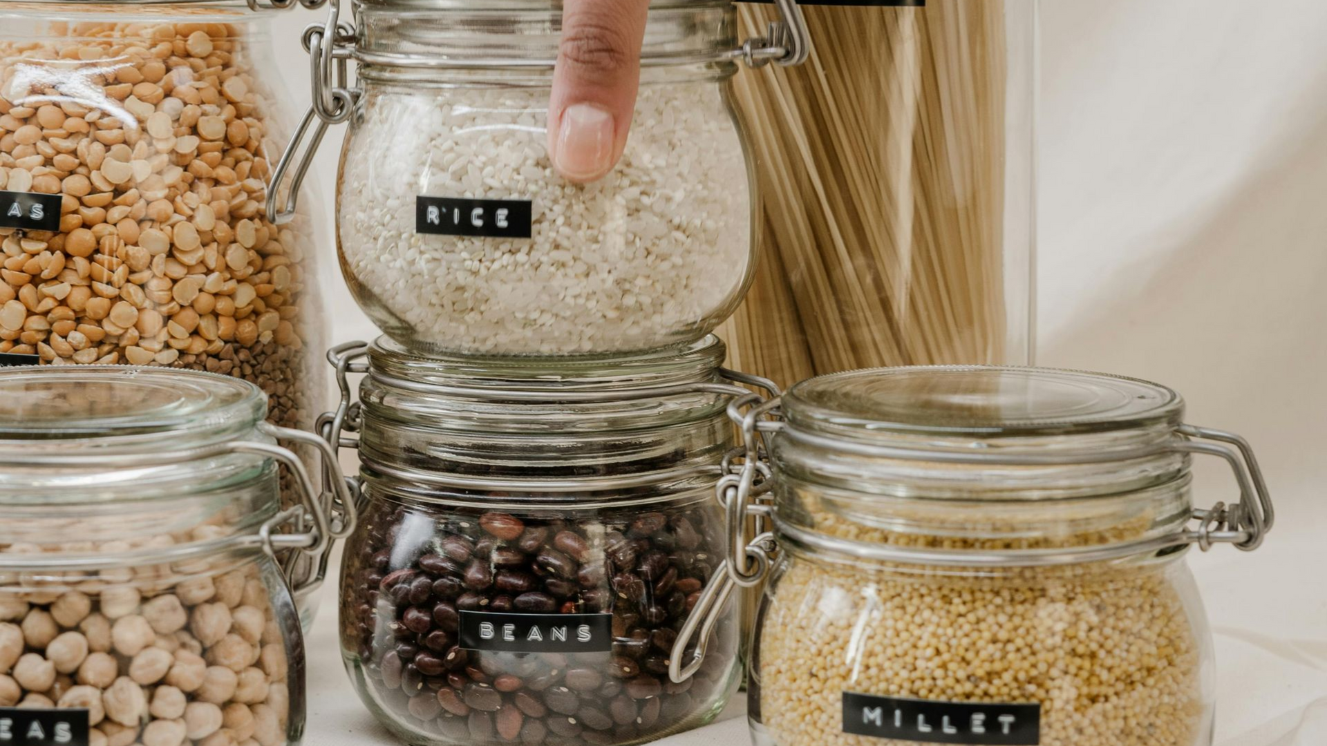 Close-up of labeled glass jars filled with grains, beans, and pasta in a pantry, with a hand pointing at a jar of rice.