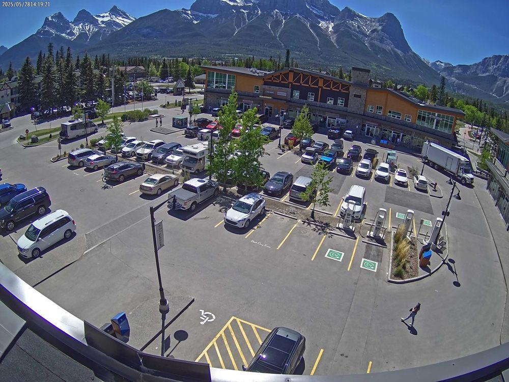 Mountain parking lot beside a lodge, with parked cars and snowcapped peaks in the background.