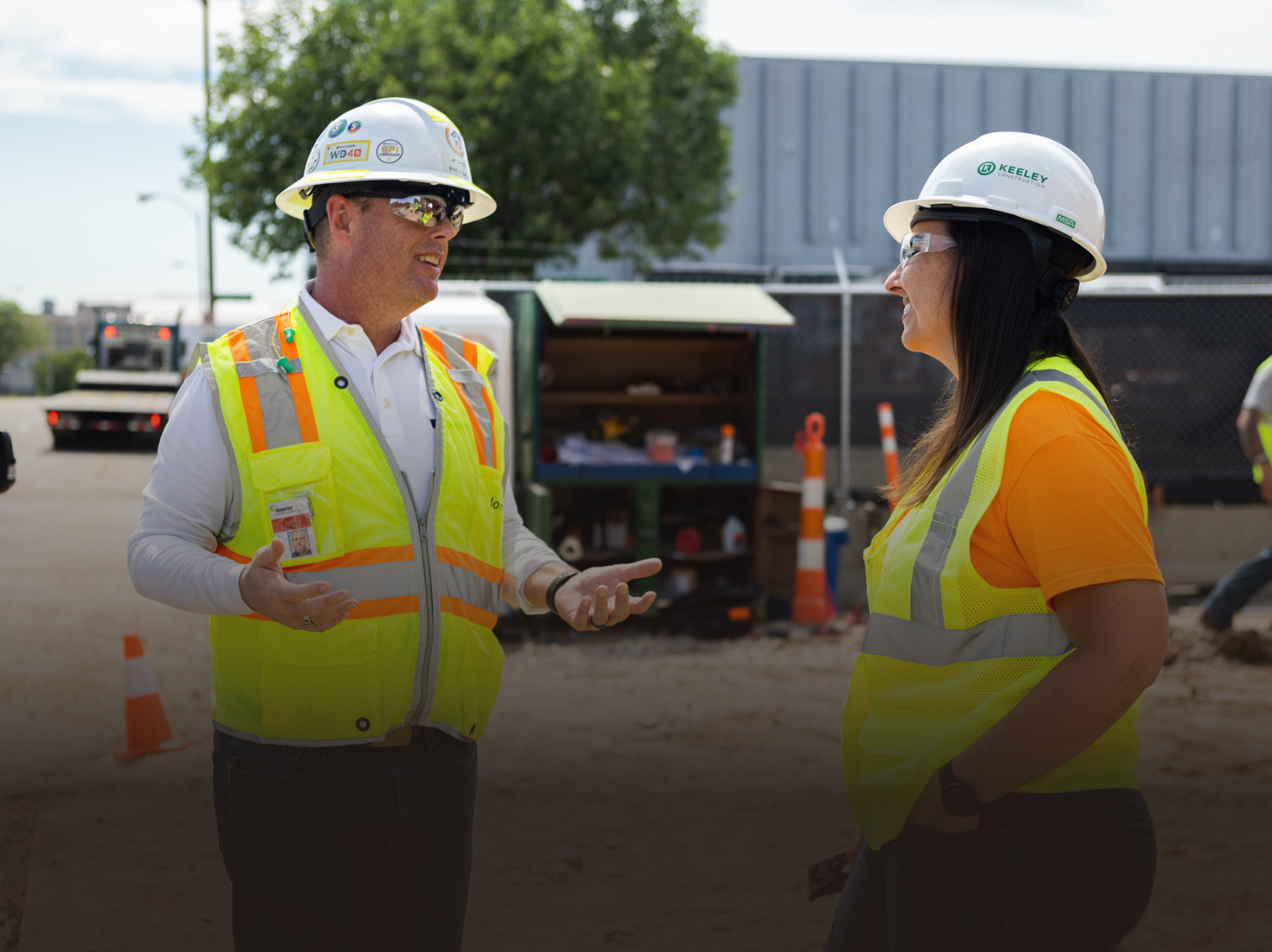 A man and a woman wearing hard hats and safety vests are talking to each other.