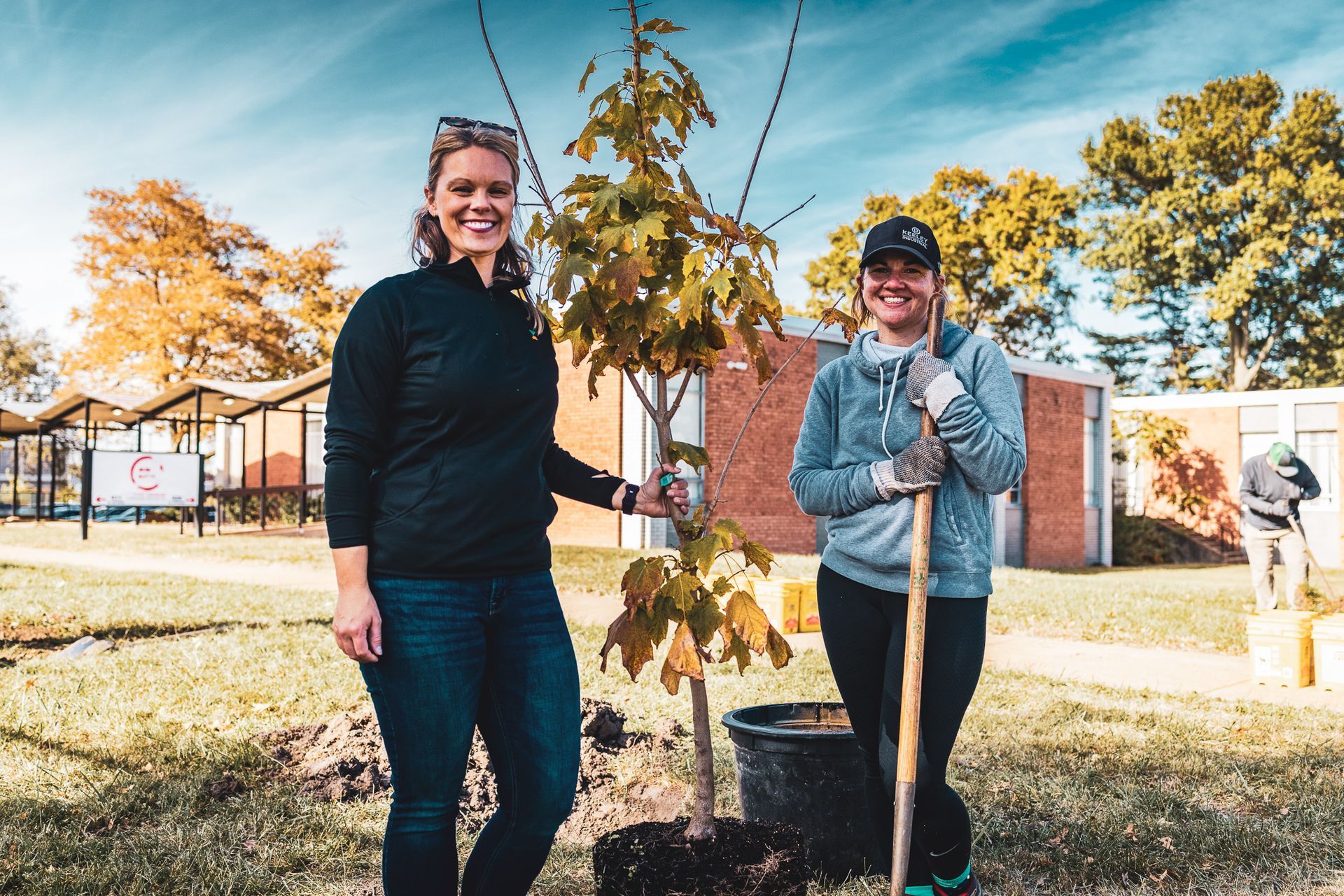 Two women are standing next to a tree in a pot.