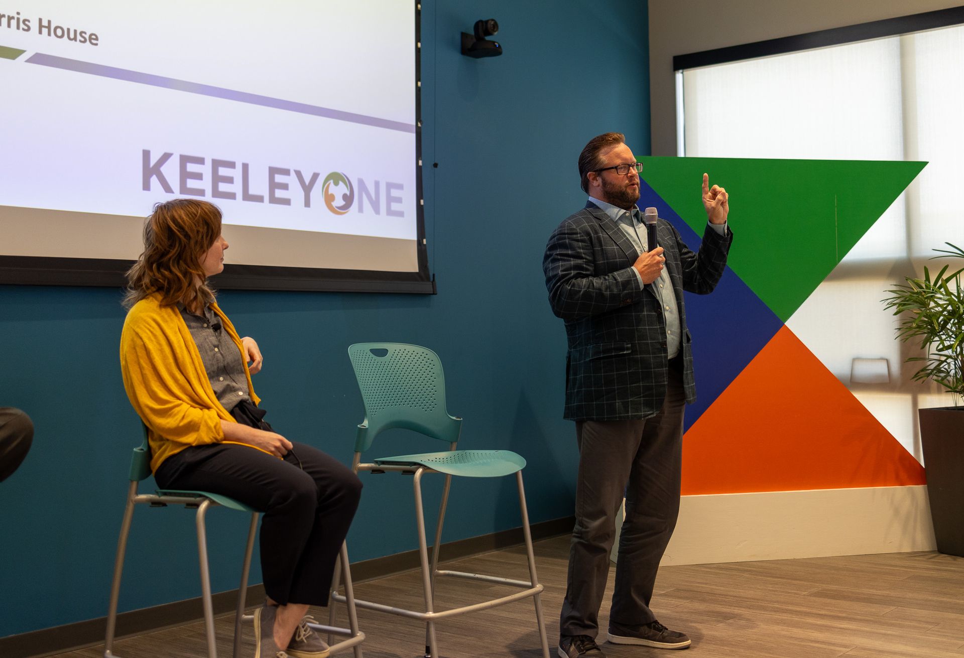 A man is giving a presentation to a group of people while a woman sits in a chair.