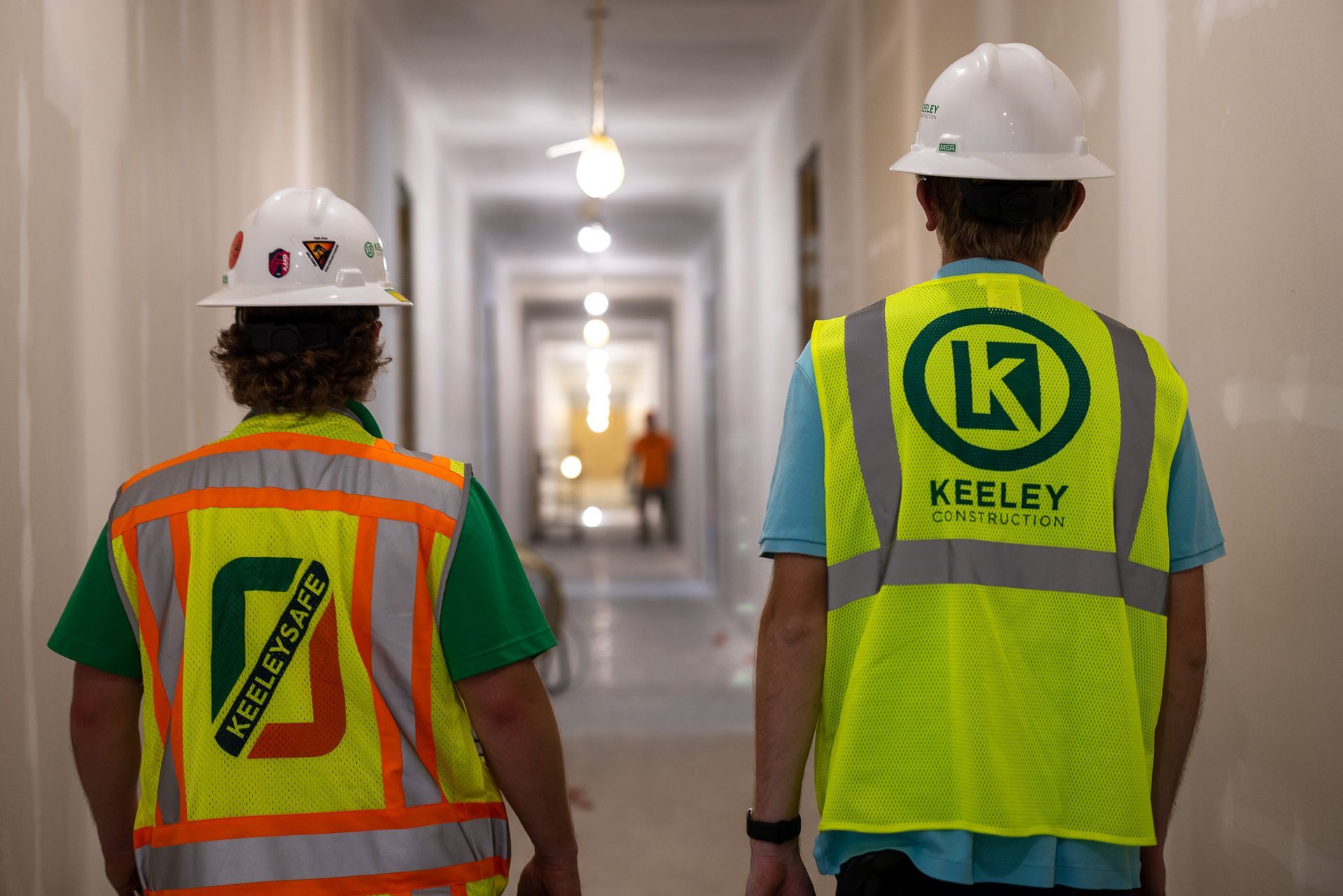 Two construction workers wearing hard hats and safety vests are walking down a hallway.