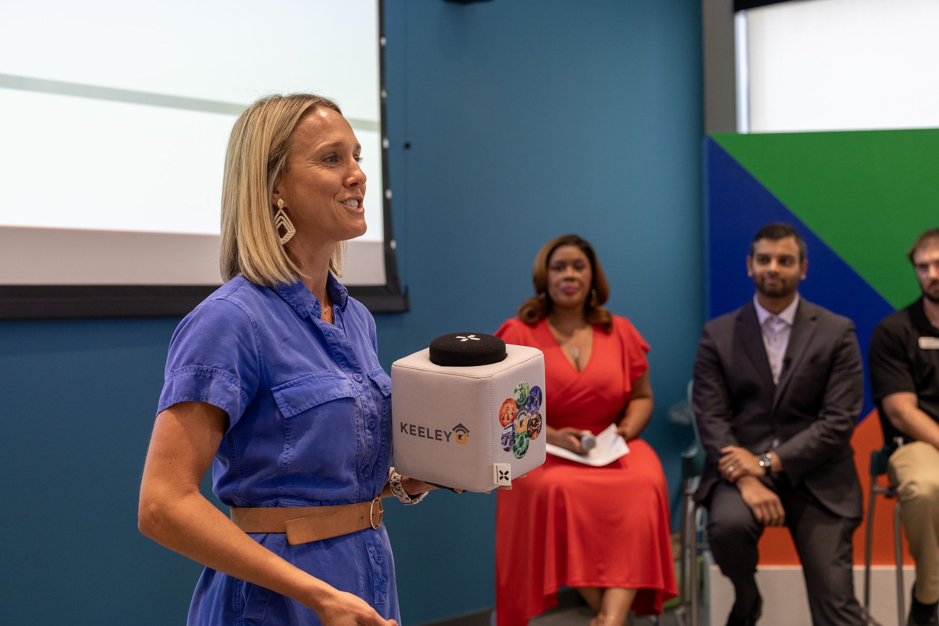 A woman is holding a cube in front of a group of people sitting in chairs.