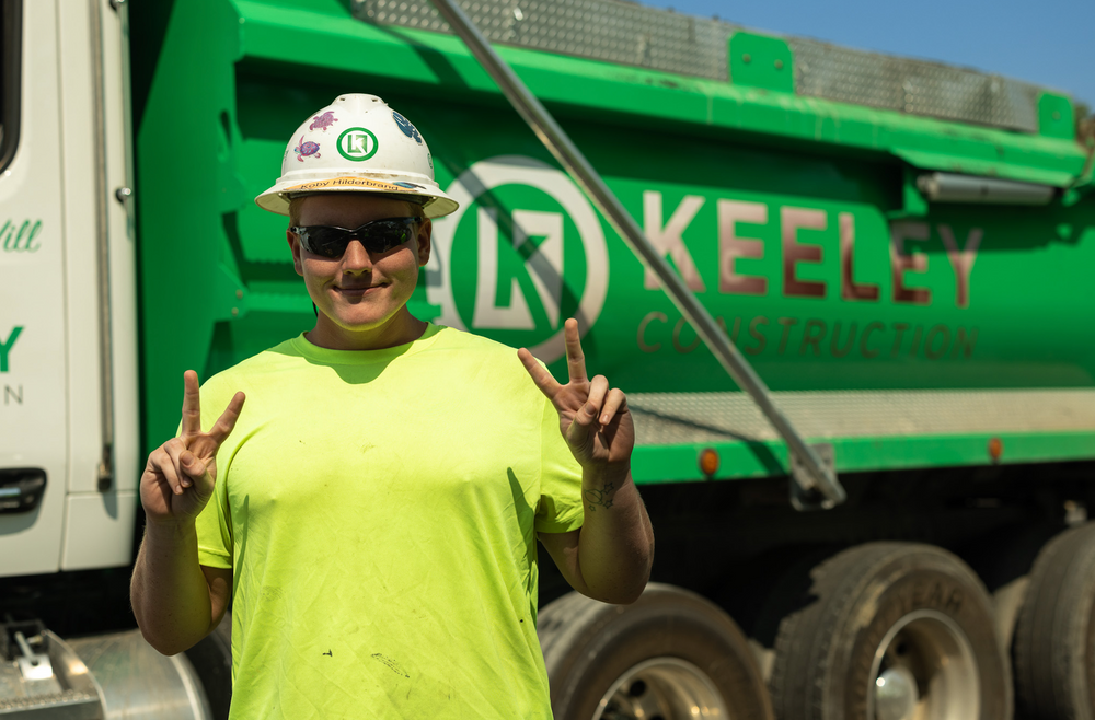 A man wearing a hard hat and sunglasses is standing in front of a green dump truck.