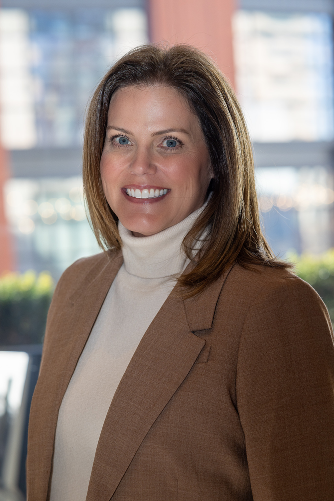 A woman in a brown jacket and white turtleneck is smiling for the camera.