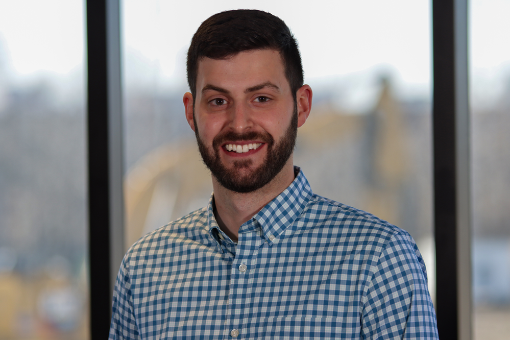 A man with a beard is wearing a blue and white plaid shirt and smiling.