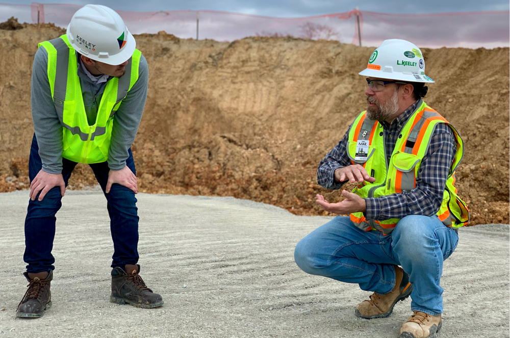 Two construction workers wearing hard hats and safety vests are kneeling down on the ground.
