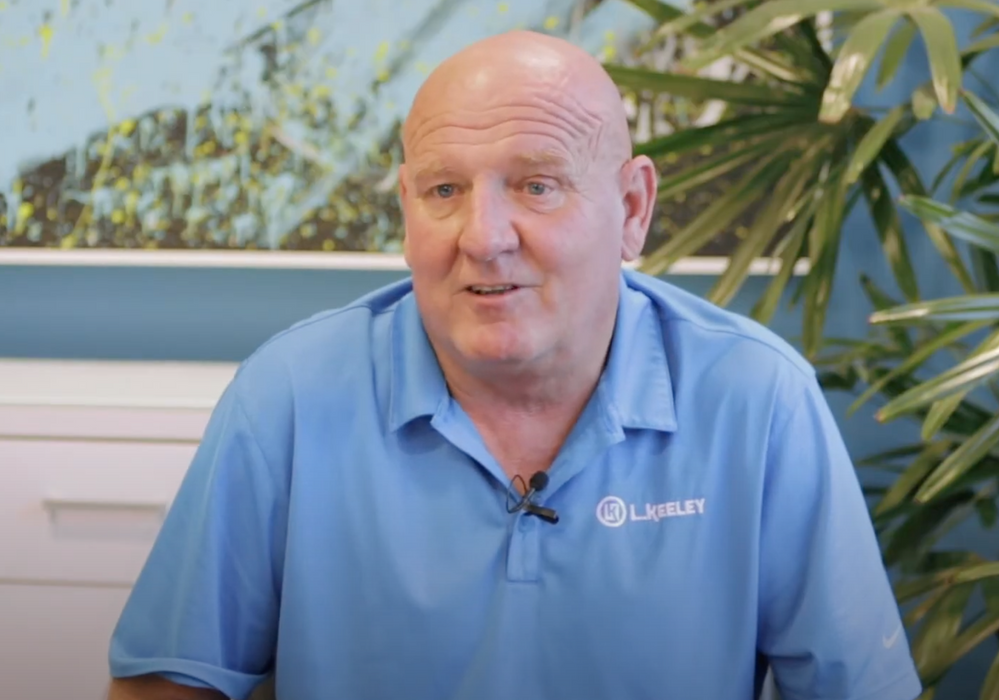 A bald man in a blue shirt is sitting in front of a plant.