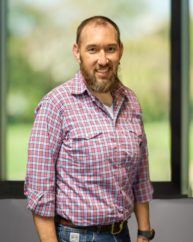 A man with a beard wearing a plaid shirt and jeans is standing in front of a window.