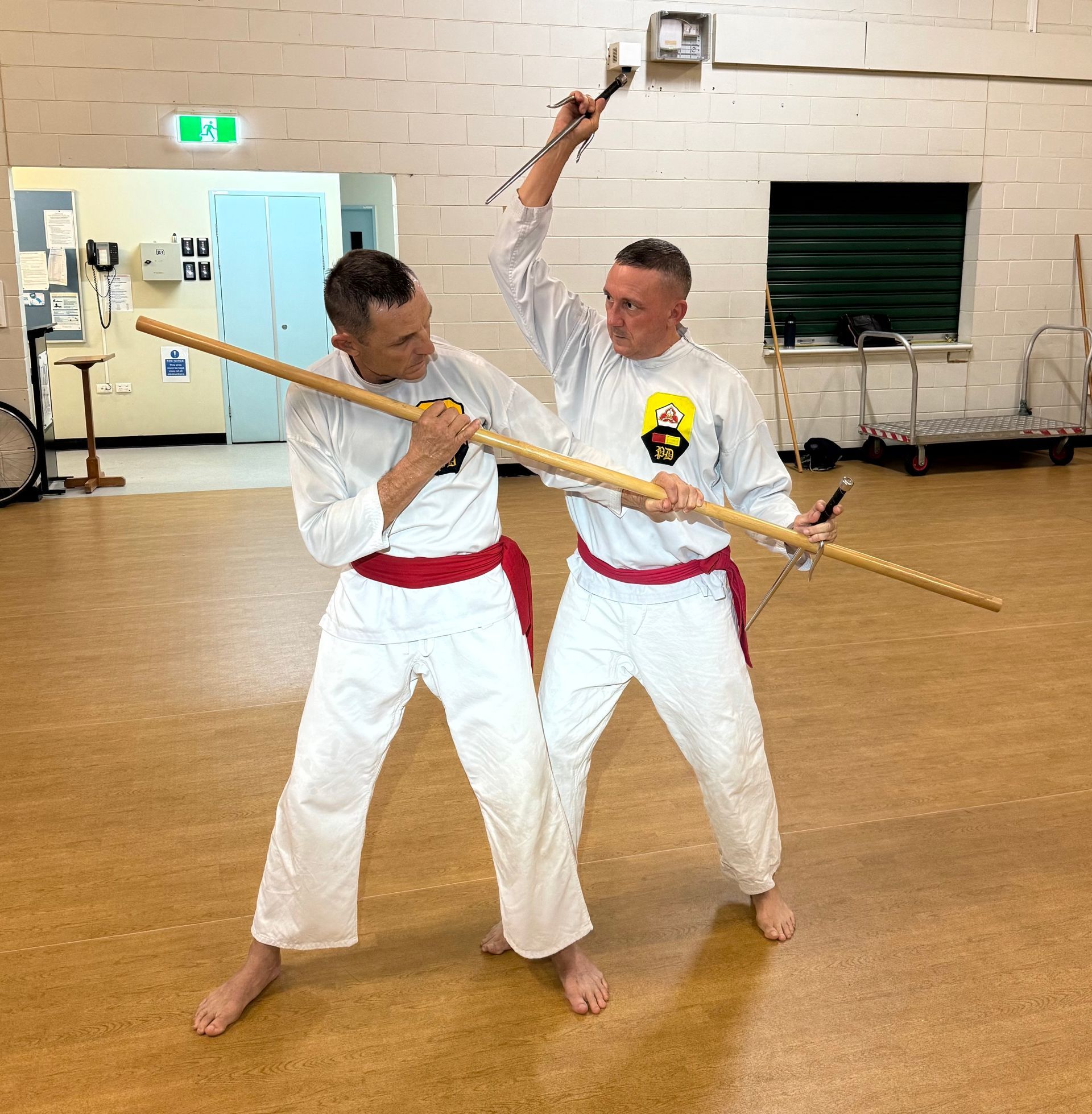 Two men in white karate uniforms are holding sticks in a room