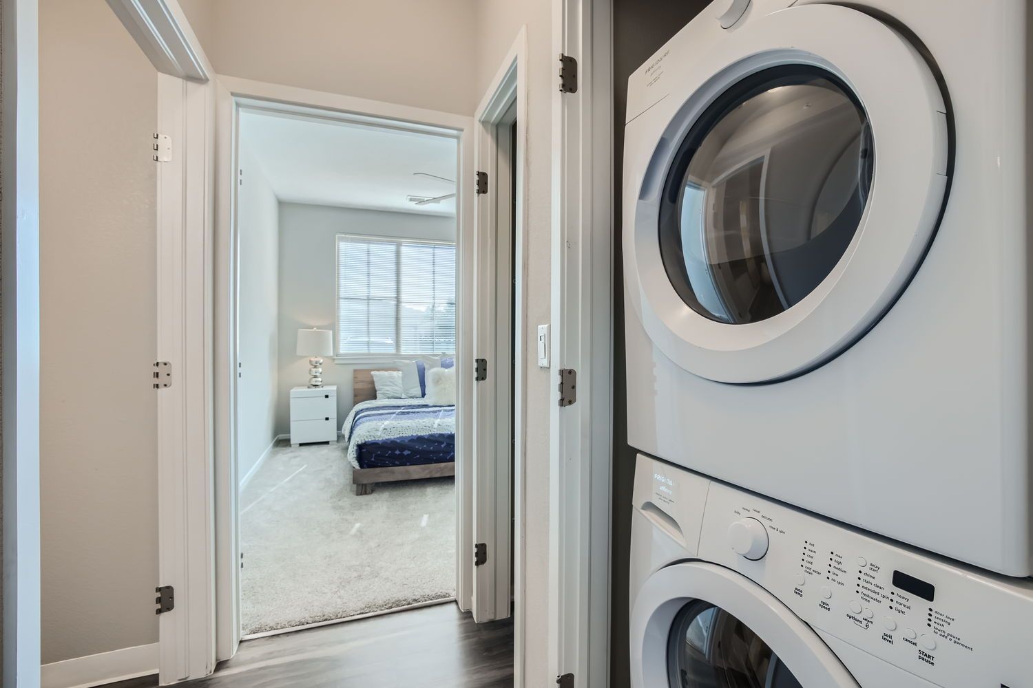 Stacked washer/dryer on the right; doorway opens to a bedroom with a bed and nightstand.
