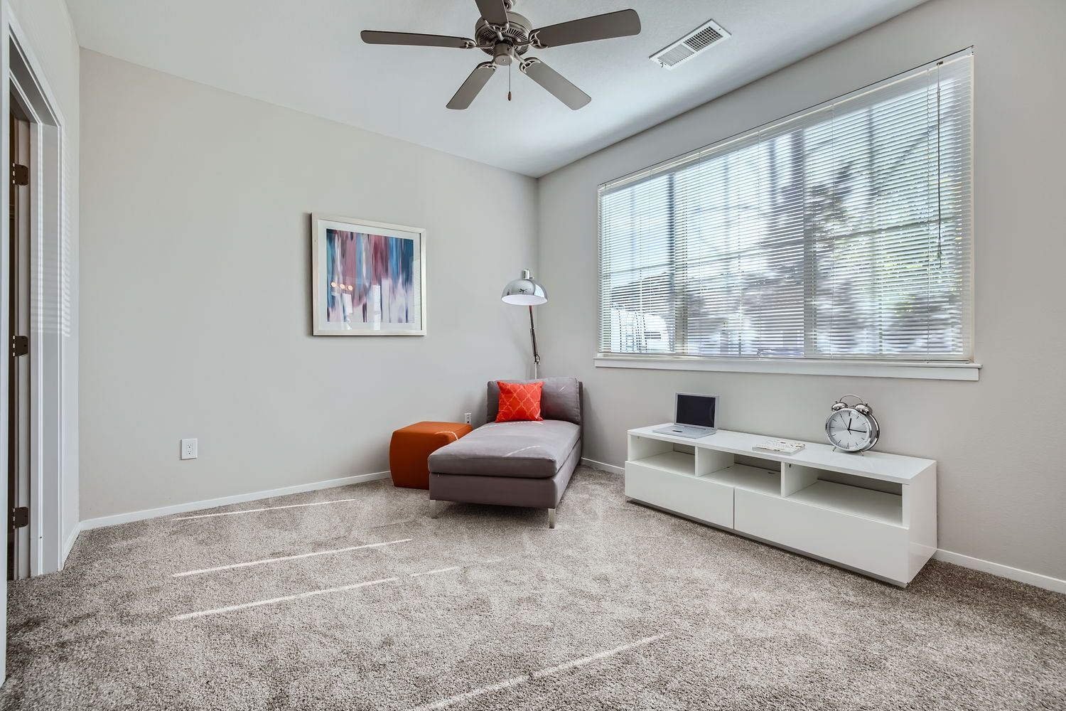 Living room with a gray chaise, orange pillow, white media console, and a large window with blinds.