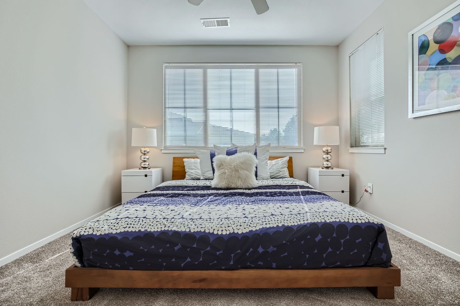 Bedroom with a low wooden platform bed, white nightstands, and two lamps by large windows with blinds.