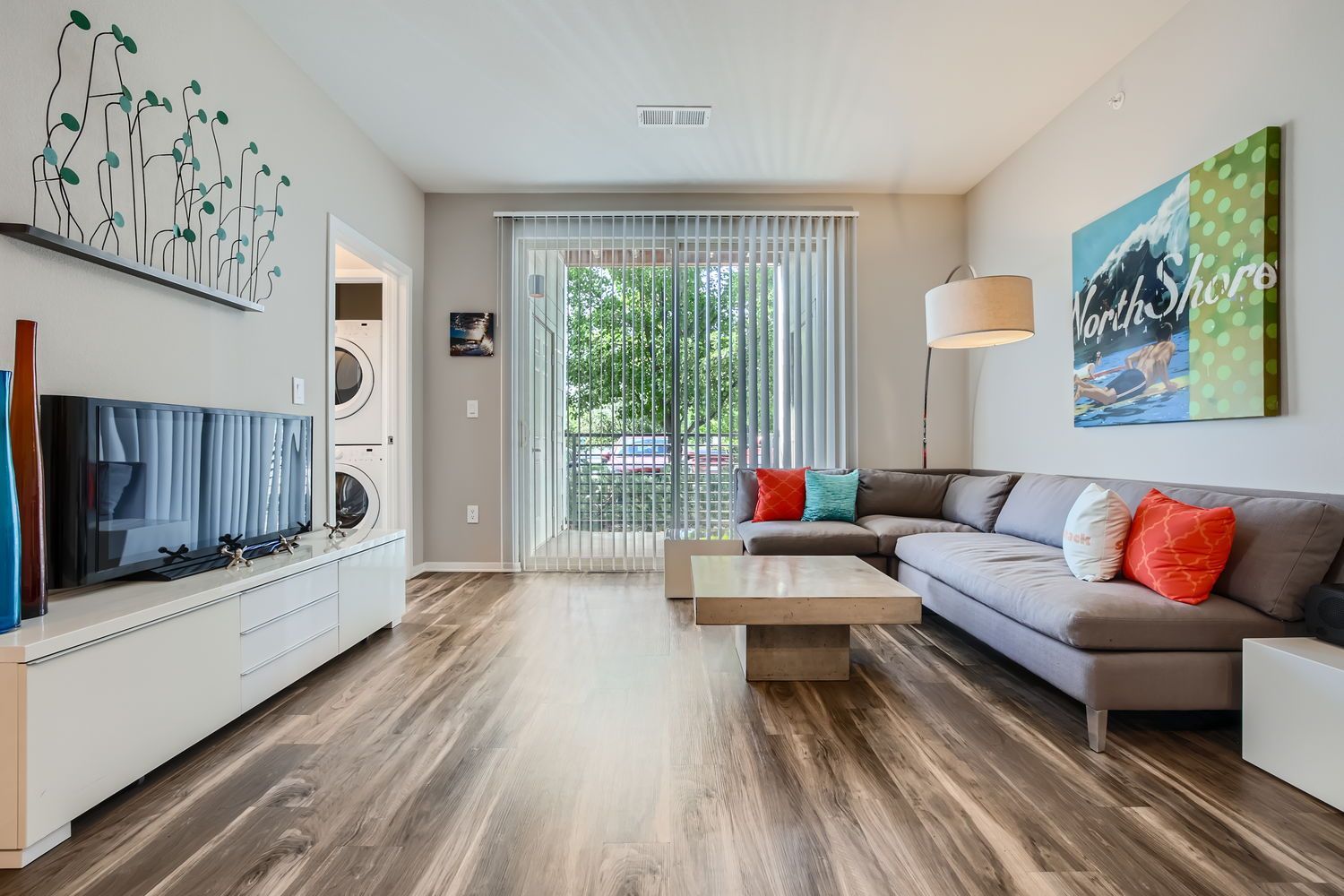 Living room in a modern apartment with a gray sectional sofa, coffee table, and sliding glass door to a balcony.