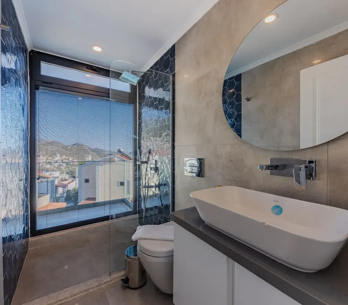 Modern bathroom with shower, toilet, wall-mounted sink, and round mirror beside a large window view