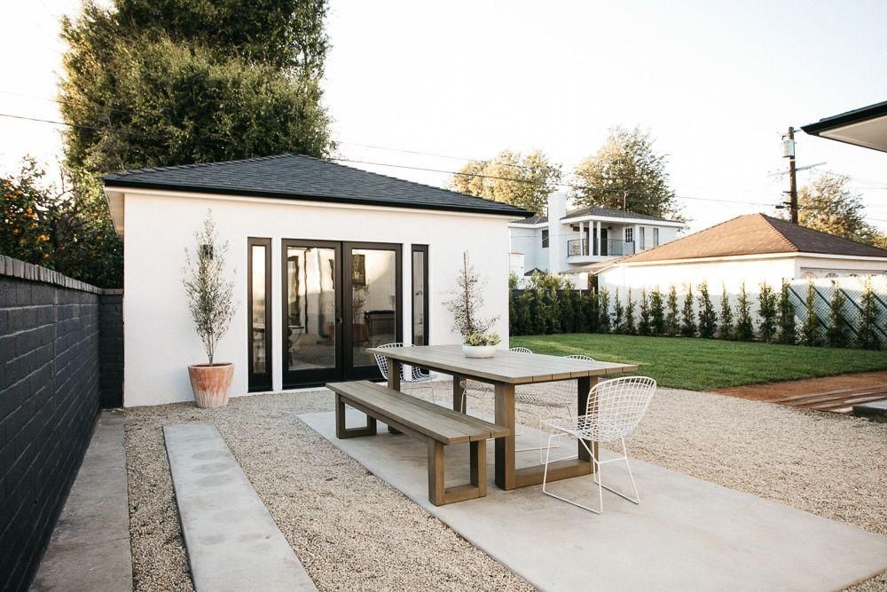 Modern backyard patio with a white garden studio, picnic table, gravel, and potted plants