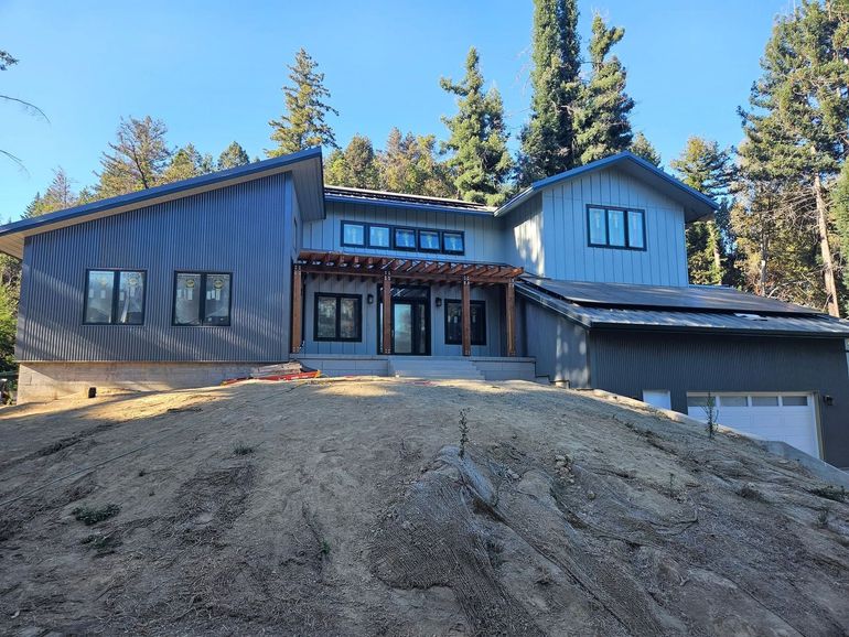 Modern dark-blue house under construction on a dirt hill, with forested trees in the background.