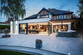Modern two-story house with open patio, warm interior lights, and outdoor seating at dusk
