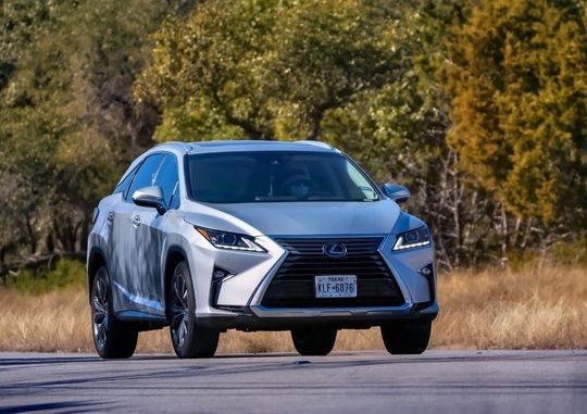 A silver lexus rx is driving down a road next to a field.