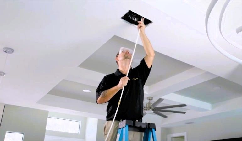 Man cleaning a ceiling vent, wearing goggles and gloves, performing duct sanitizing.