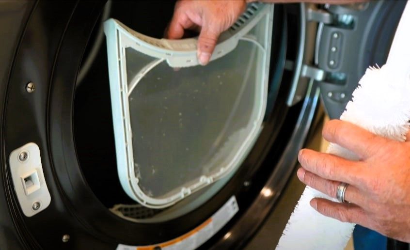 Hand holding a lint filter in front of an open clothes dryer.