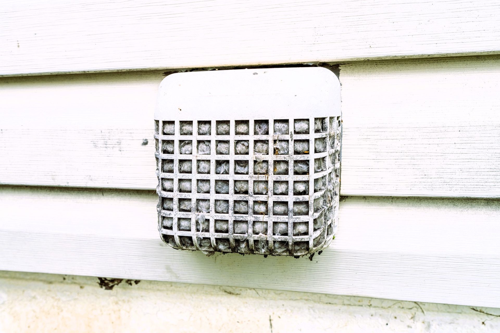 Dusty exterior dryer vent cover on a white horizontal siding wall.