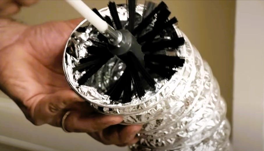 Person cleaning dryer vent with a brush inserted in a silver duct pipe.