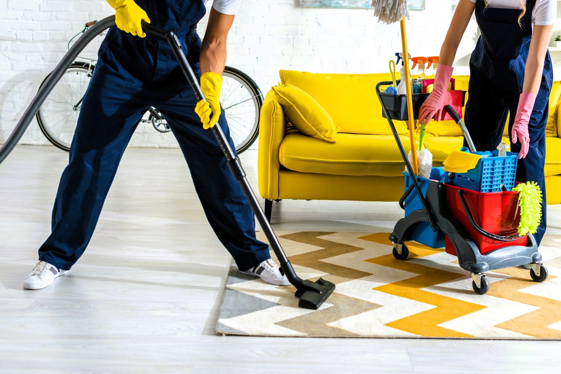 Two people in overalls cleaning a room with a vacuum cleaner and a cart of supplies near a yellow sofa.