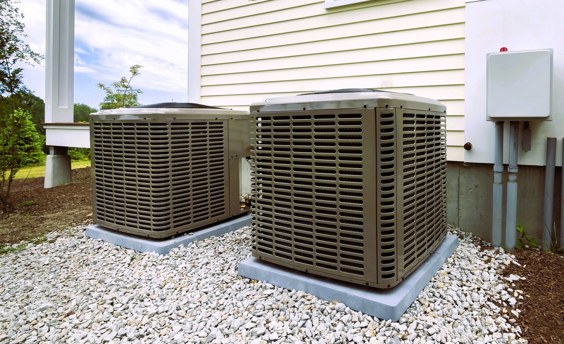 two outdoor HVAC units on gravel next to a house with visible electrical connections