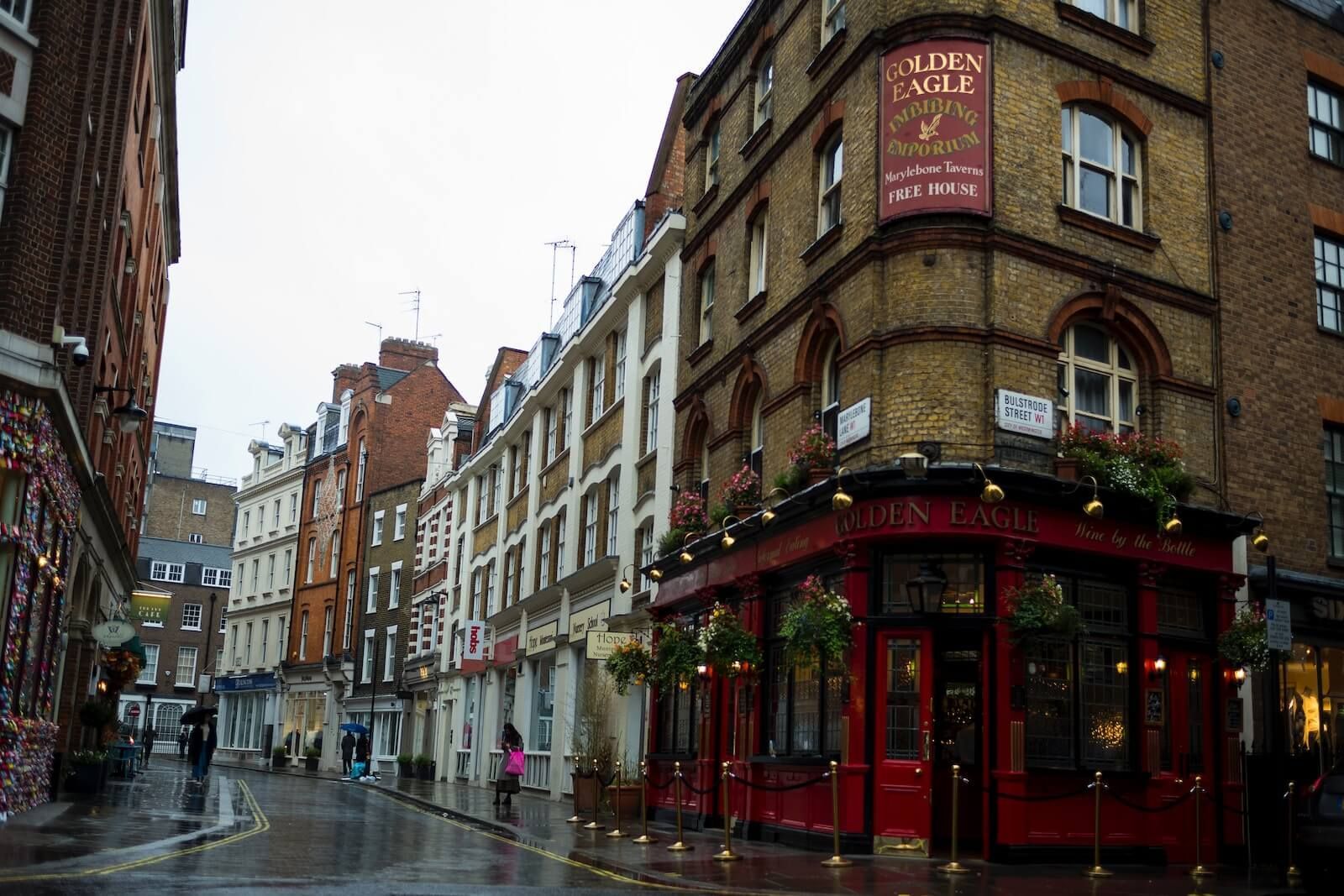 Street view of London buildings, including The Eagle pub with red exterior and sign. Wet cobblestone street.